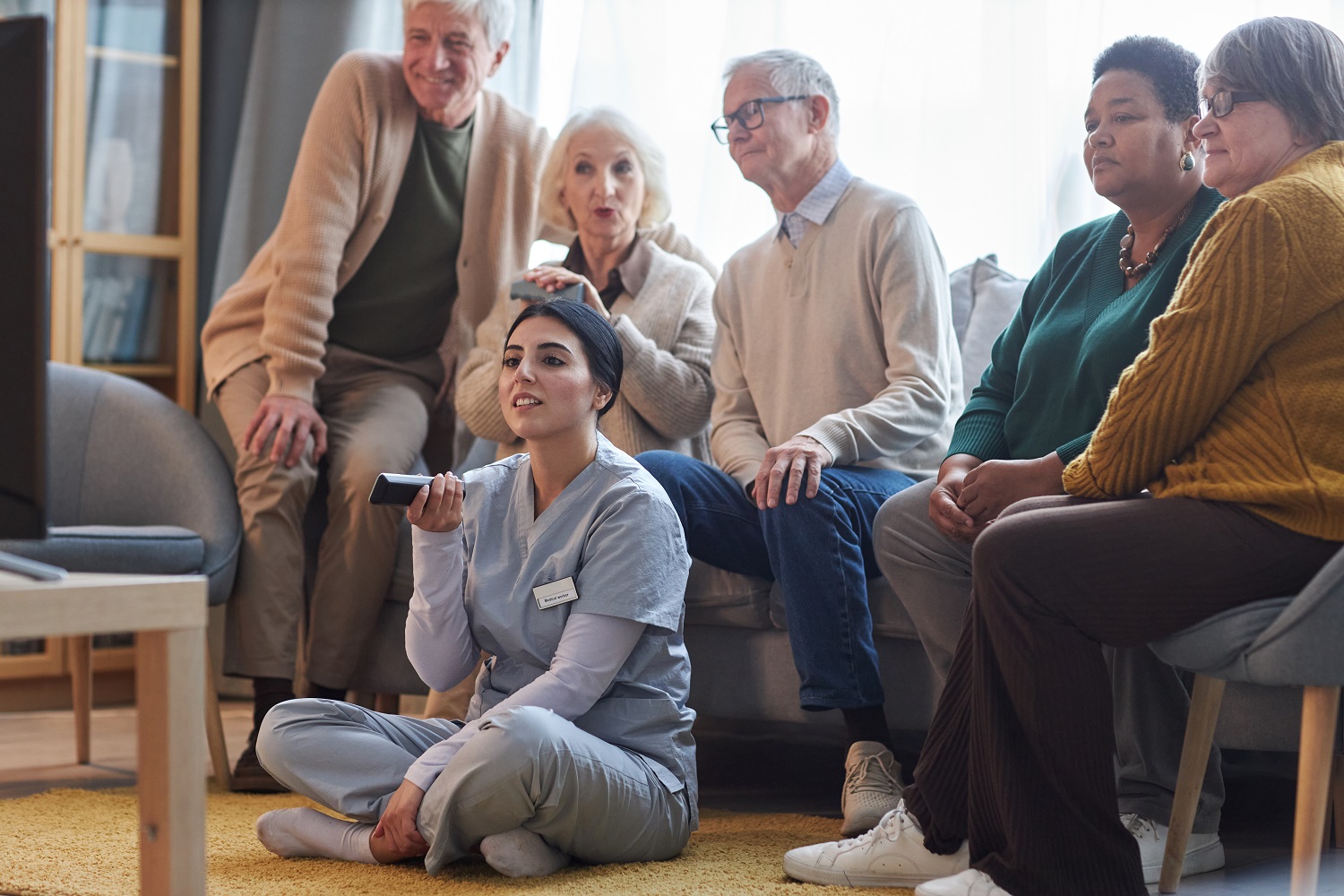 Diverse group of senior people watching TV in retirement home together with young nurse holding remote